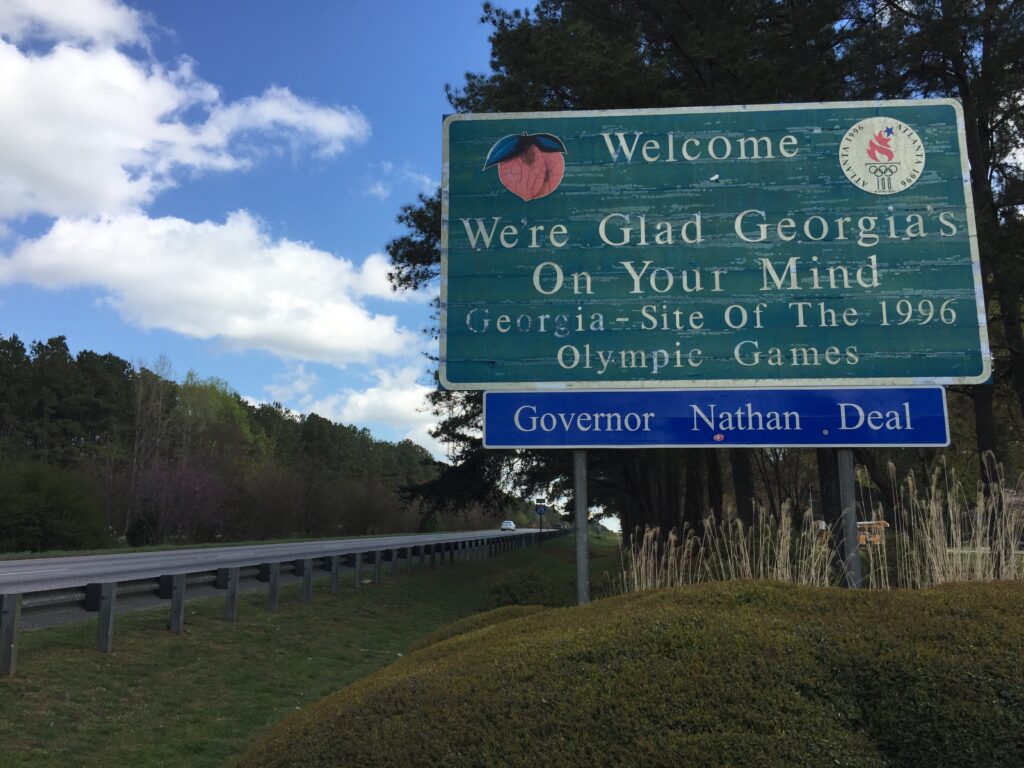 A photograph of a road sign that says "Welcome We're Glad Georgia's On Your Mind Georgia - Site of the 1996 Olympic Games"