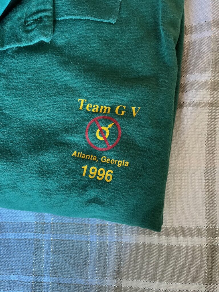 A green polo shit with a little logo printed on it in yellow letters that says "Team GV, Atlanta, Georgia 1996" and has a little symbol for male with a red circle and cross through it. 