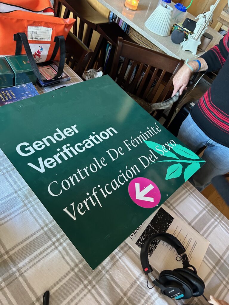 A green sign that says "Gender Verification, Conrole de Feminite, Verificacion del Sexo" on it and a pink arrow pointing to the left.