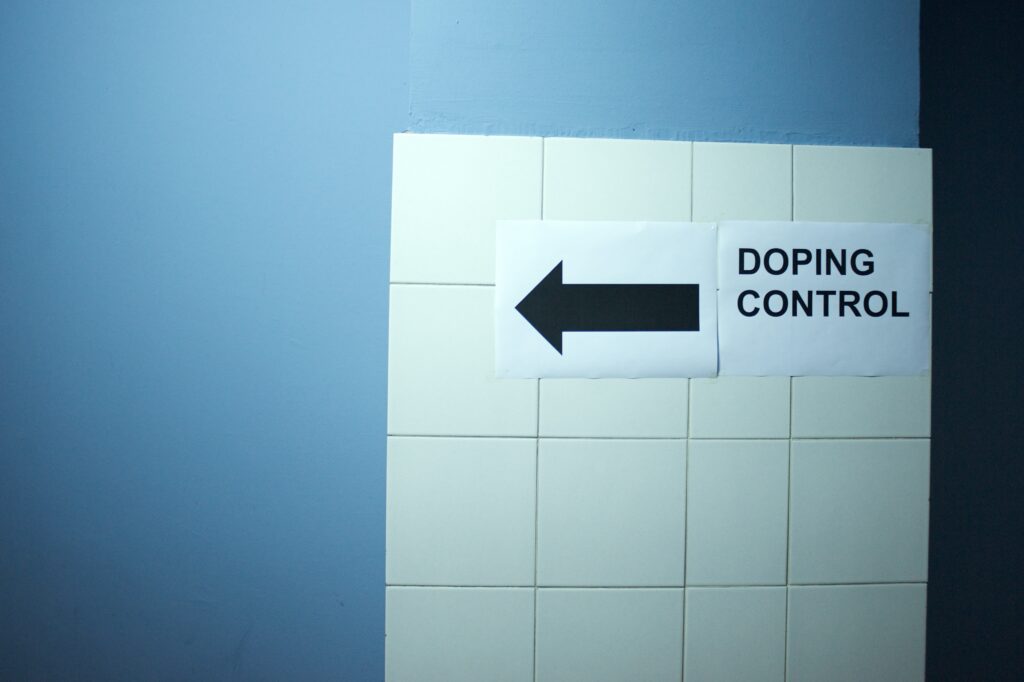 A blue and white wall with a paper sign taped up that says "doping control" with a black arrow pointing left